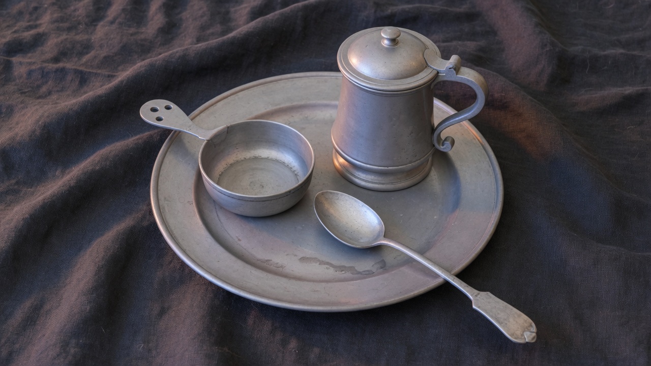 Unlabeled pewter plate, porringer, lidded tankard, and spoon on dark linen